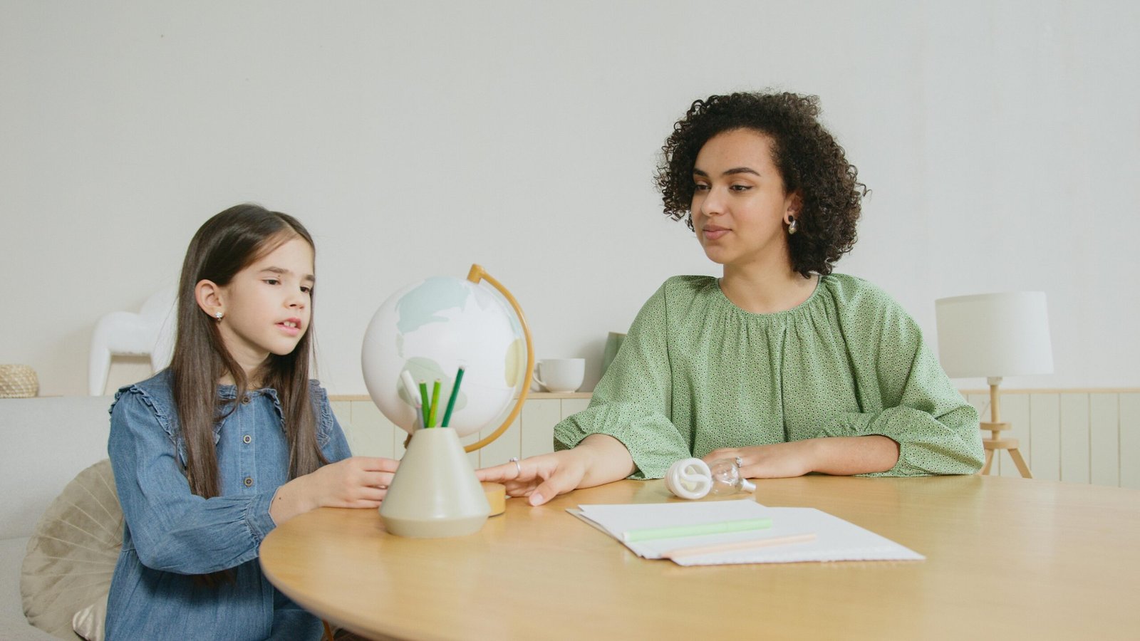 African American woman teaching caucasian girl with a globe in a classroom setting.